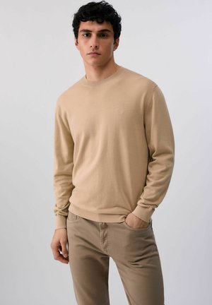 Joven con cabello rizado oscuro, vestido con suéter beige y pantalones a juego, de pie con una mano en el bolsillo contra un fondo liso.