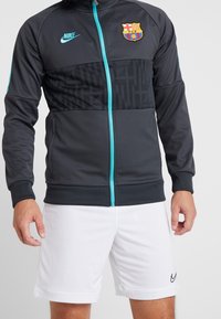 Ciemnoszara kurtka z suwakiem Nike z turkusowymi akcentami, z logo Barcelony i teksturowanym panelem. Nosi ją osoba w białych szortach.