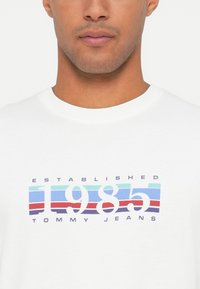 Άνδρας που φοράει λευκό T-shirt με πολυχρωμο κείμενο "1985" και "Tommy Jeans Established" στο στήθος.