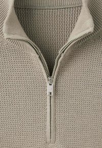 Gros plan d'un pull beige en maille avec une fermeture éclair argentée partiellement ouverte et un col côtelé.