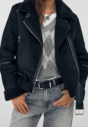 Person trägt schwarze Lammfelljacke, grauen Argyle-Pullover, hell gewaschene Jeans mit schwarzem Gürtel und silberne Halskette mit Herzanhängern.