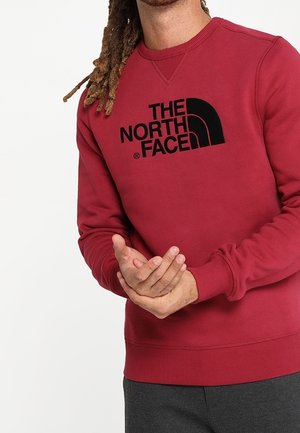 Sudadera marrón con cuello redondo y puños acanalados, con un gran logo negro de "The North Face" en la parte frontal.