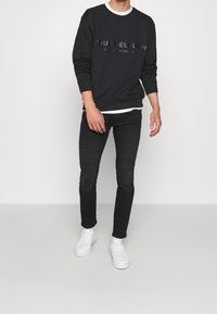 Černá mikina s tónovým logem "True Religion", v kombinaci s černými džíny a bílými teniskami. Mikina má kulatý výstřih a žebrované manžety.