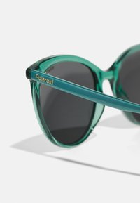 Ochelari de soare tip cat-eye verzi, cu ramă translucidă și lentile închise. Prezintă un logo metallic auriu "Polaroid" pe templu. Textură netedă.