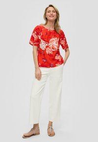 Rote Bluse mit floralem Muster, kurzen Ärmeln und rundem Ausschnitt. Kombiniert mit weißen, weit geschnittenen Hosen und silbernen Sandalen. Glattes Material, lebendiges Druckmuster.
