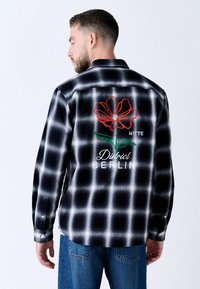 Mustmustruuduline särk, millel on punased lilleämbroidery, tekst "MITTE" ja "District BERLIN", pikad varrukad ja ümar hem.}}