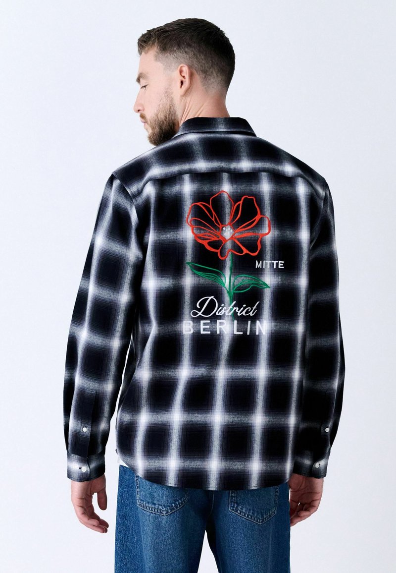 Camicia a scacchi bianco e nero con ricamo di fiori rossi, testo "MITTE" e "District BERLIN", con maniche lunghe e orlo arrotondato.