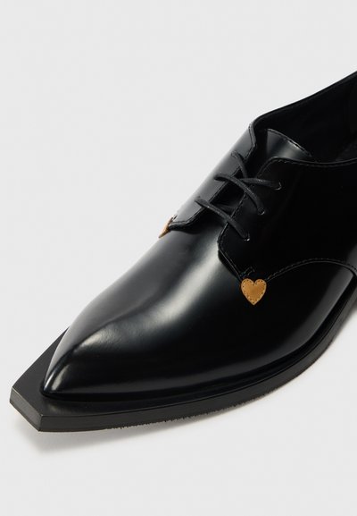 Fekete hegyes bőrcipő, egyedi szögletes orral és kis barna szív alakú díszekkel a fűzőknél, fehér háttér előtt.