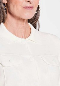 Witte gebreide cardigan met een omgeslagen kraag en twee borstzakken. Bevat ronde knopen en een textuurstof. Oorbellen zijn zichtbaar op de afbeelding.