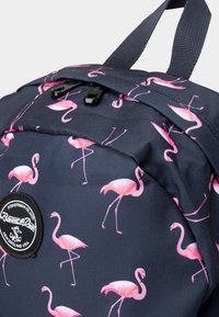 Zaino navy con motivo ripetuto di fenicotteri rosa, dotato di chiusura superiore a zip, accessori neri e un'etichetta rotonda con logo.