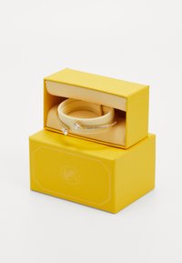 Bracciale d'oro con due pietre trasparenti a forma di cuore e piccoli cristalli, esposto all'interno di una scatola di gioielli gialla su uno sfondo bianco.