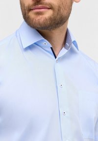Camicia azzurro chiaro con colletto decorato, design a bottoni e taschino sul lato sinistro del petto, realizzata in un tessuto morbido. Presenta bottoni bianchi.