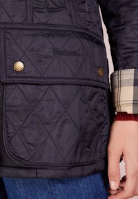 Detailní záběr na kapsu námořnického prošívaného saka s logem Barbour, bronzovými cvoky, stočeným kostkovaným manžetou a rukou spočívající vedle kapsy.