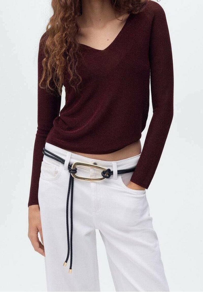 Femme portant un haut moulant bordeaux foncé à manches longues et col en V, rentré dans un pantalon blanc taille haute avec une ceinture corde noire et une boucle dorée.