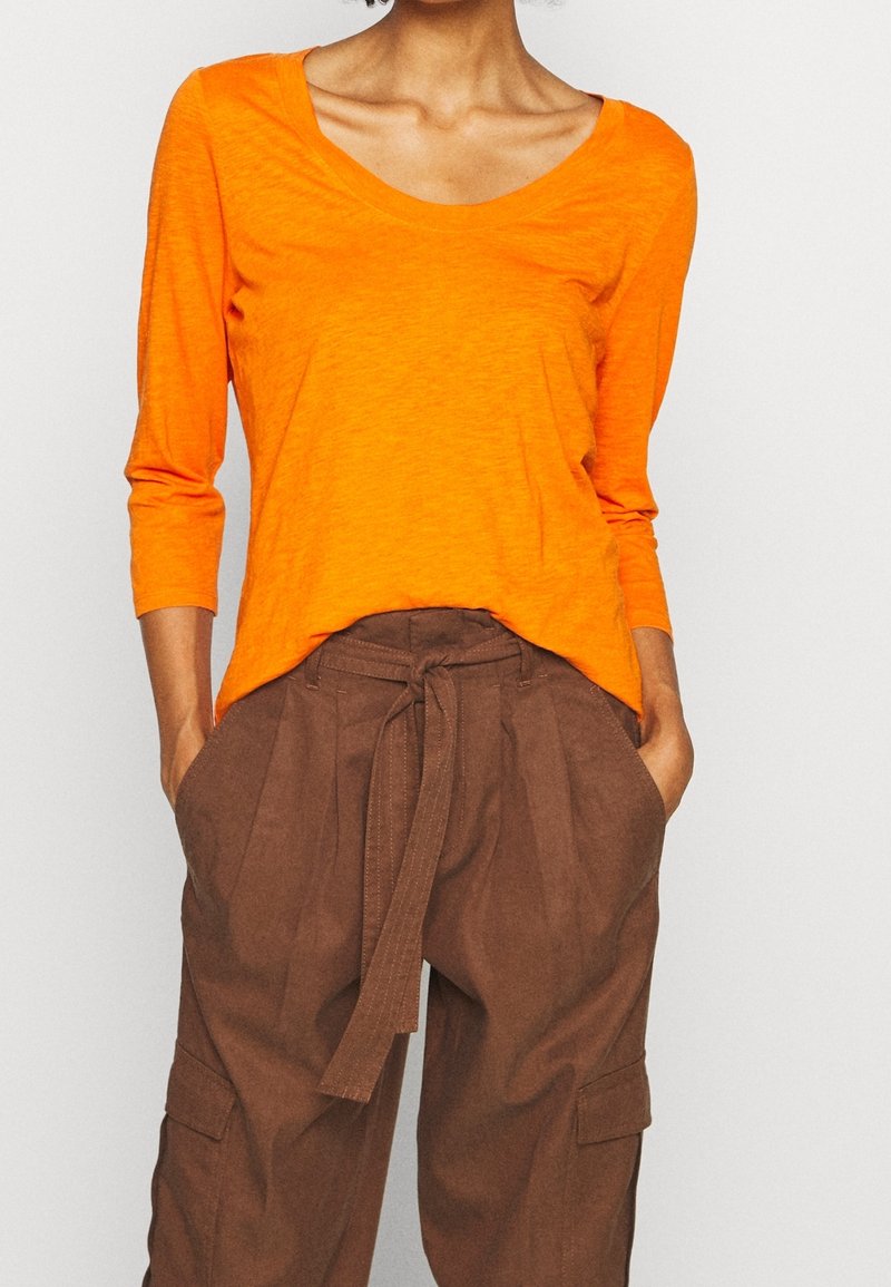 Maglia a maniche lunghe, con scollo a barca in tessuto arancione brillante, abbinata a pantaloni marroni a vita alta con cintura annodata e tasche laterali.