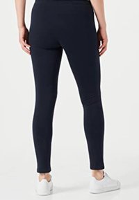 Sötétkék leggings, rugalmas anyagból készült, testhezálló kialakítással, sima textúrával és bokáig érő hosszúsággal, fehér lábbelivel párosítva.