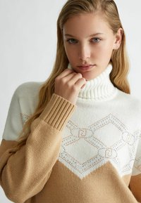 Maglione a maniche lunghe con motivo crema e tan, collo alto e polsini a coste, caratterizzato da design geometrici e sottili decorazioni. Texture in maglia morbida.