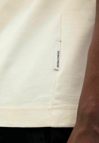 Crème-kleurig t-shirt van katoen, met een zijtag met de merknaam. Heeft een gladde textuur en een eenvoudig ontwerp.