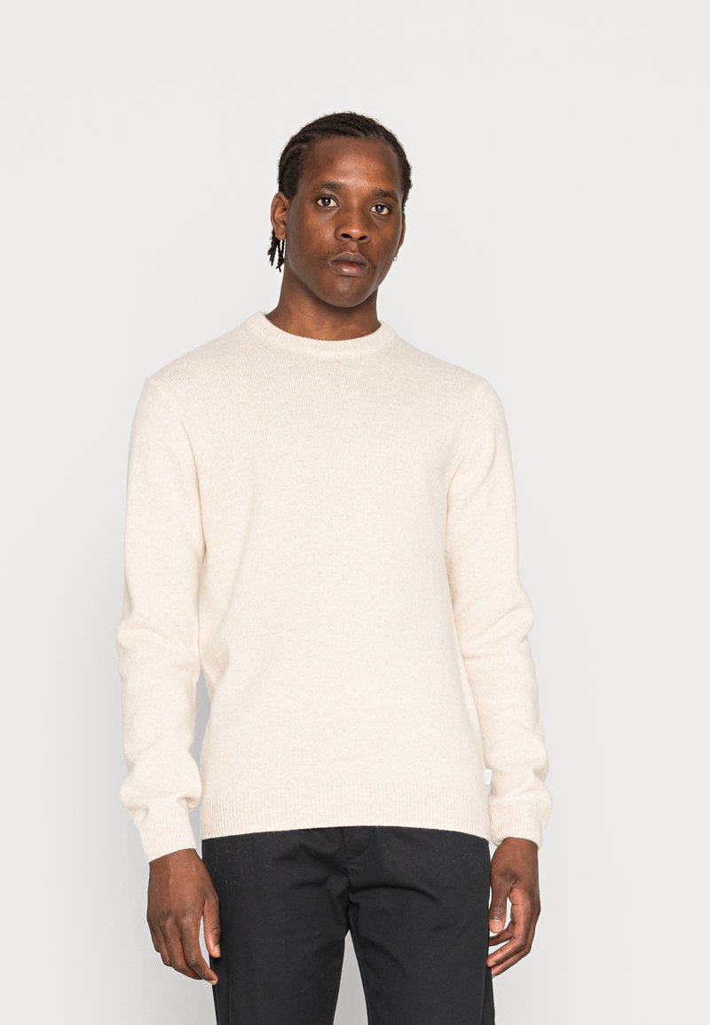 Beige Pullover mit Rundhalsausschnitt, langen Ärmeln und gerippten Bündchen. Weiche Textur, hergestellt aus einem Strickstoff. Kombiniert mit dunklen Hosen.