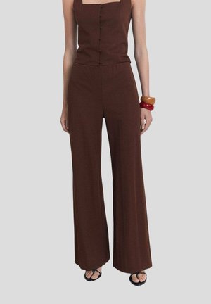 Donna che indossa un top marrone senza maniche con bottoni e pantaloni larghi abbinati, sandali neri e braccialetti spessi arancioni e rossi al polso destro.