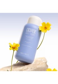 Coco & Eve Boost Therapy volymgivande balsam i en mjuk blå flaska med vitt lock, placerad på en sten med gula blommor.