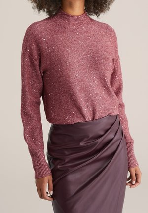 Femme portant un pull mauve scintillant en maille, associé à une jupe taille haute en similicuir bordeaux foncé avec un détail drapé.