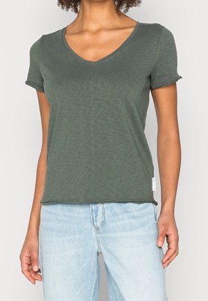 Kobieta w dopasowanej ciemnozielonej koszulce z krótkim rękawem i dekoltem w serek oraz jasnoniebieskich, wysokich dżinsach, stojąca na jednolitym tle.