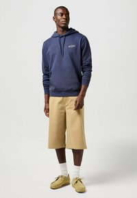 Felpa navy con tasca frontale e cordino, abbinata a pantaloni corti beige al ginocchio e scarpe color carne. Il modello è in piedi davanti a uno sfondo neutro.