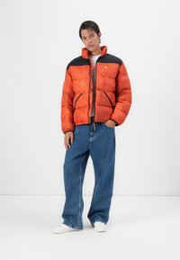 La veste en duvet orange présente un design texturé, des accents noirs aux épaules et une fermeture éclair à l'avant. Elle est associée à un jean en denim bleu ample et des chaussures blanches.