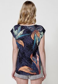 Chemise à manches courtes avec un motif floral sombre et des feuilles vibrantes en orange, vert et bleu, en tissu léger.