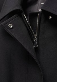 Gros plan d’un col de manteau noir avec une fermeture éclair argentée partiellement ouverte et deux boutons-pression métalliques sur les revers.