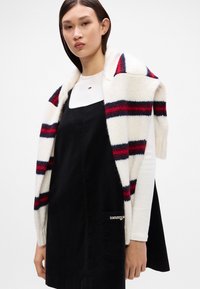 Ριγέ πλεκτό κασκόλ σε κρεμ χρώμα με κόκκινες και ναυτικές λεπτομέρειες πάνω από ένα μαύρο τζιν φόρεμα με τετράγωνη λαιμόκοψη και μία πλαϊνή τσέπη.