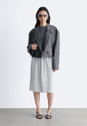 Femme portant un blazer gris surdimensionné, un haut sombre, une jupe grise claire longueur genou, des talons noirs à lanières, des lunettes de soleil et des boucles d'oreilles en créoles, debout sur un sol gris.