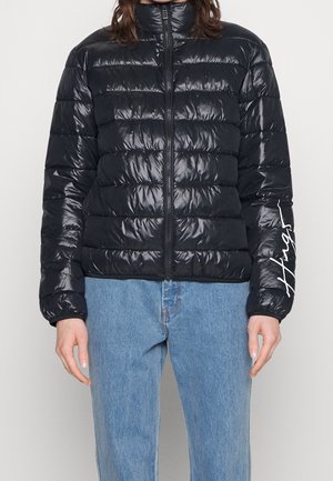 Schwarze glänzende gesteppte Steppjacke mit Frontreißverschluss und weißer Schrift am linken Ärmel, kombiniert mit hellblauen Jeans.