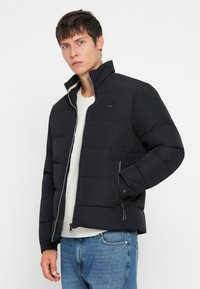 Giovane uomo che indossa un giubbotto imbottito nero sopra un maglione bianco e jeans blu, con una mano nella tasca della giacca, su uno sfondo neutro.