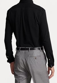 Hombre con una camisa negra de manga larga metida dentro de pantalones de vestir gris claro, con un cinturón negro, visto desde atrás.