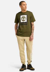 Olivově zelené tričko s černobílým logem Timberland; v kombinaci s béžovými tepláky s nápisem Timberland umístěným vertikálně.