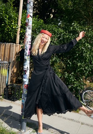Femme en robe noire et chapeau rouge posant joyeusement près d'un poteau recouvert d'autocollants sur un trottoir ensoleillé avec des buissons verts à l'arrière.