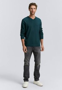 Dunkelgrüner V-Ausschnitt-Pullover mit weicher Textur, kombiniert mit grauer Jeans und weißen Turnschuhen. Der Pullover hat lange Ärmel und gerippte Bündchen.