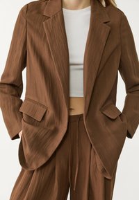 Femme portant une veste de costume marron texturée et un pantalon assorti avec un haut blanc court côtelé, les mains dans les poches, sur fond blanc.