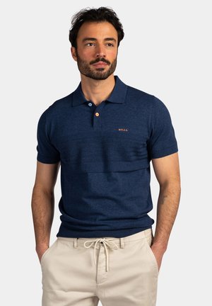 Man met baard die een marineblauw poloshirt met korte mouwen en beige broek met trekkoord draagt, staand met handen in de zakken tegen een effen achtergrond.