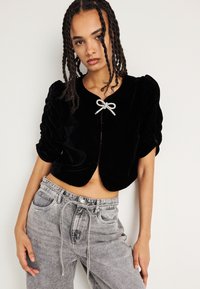 Giacca cropped in velluto nero con maniche a sbuffo, caratterizzata da un dettaglio in satin argento alla scollatura e un orlo arrotondato. Indossata con jeans grigi a vita alta.