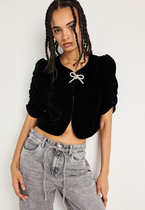 Črna žametna skrajšana jakna z napihnjenimi rokavi, ki ima srebrno pentljo na ovratniku in okrogel rob. Nosi se z sivo visokim pasom.