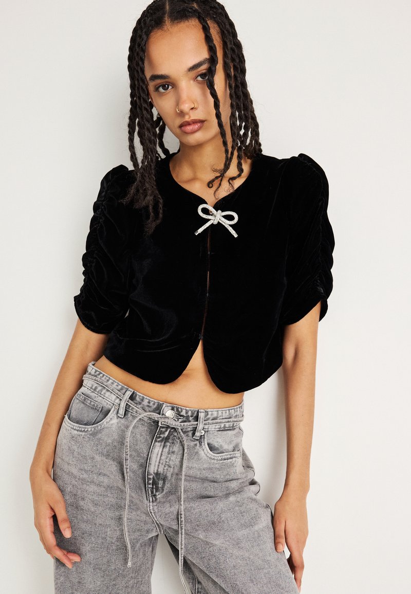 Giacca cropped in velluto nero con maniche a sbuffo, caratterizzata da un dettaglio in satin argento alla scollatura e un orlo arrotondato. Indossata con jeans grigi a vita alta.