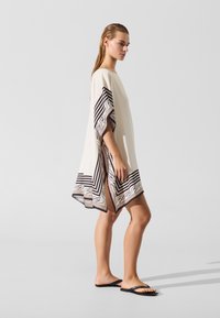 Kremasti kaftan sa širokim rukavima, s geometrijskim uzorkom u crnoj i smeđoj boji duž ruba. Model nosi crne japanke.