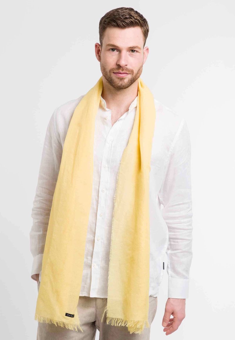 Yellow scarf made of lightweight fabric, featuring fringed edges. Worn over a white button-up shirt, with a soft, smooth texture.