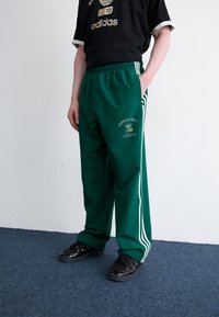 Zelene športne hlače s tremi belimi črtami, z elastičnim pasom in detajlom logotipa, v kombinaciji z črnimi športnimi copati na modrem tepihu.