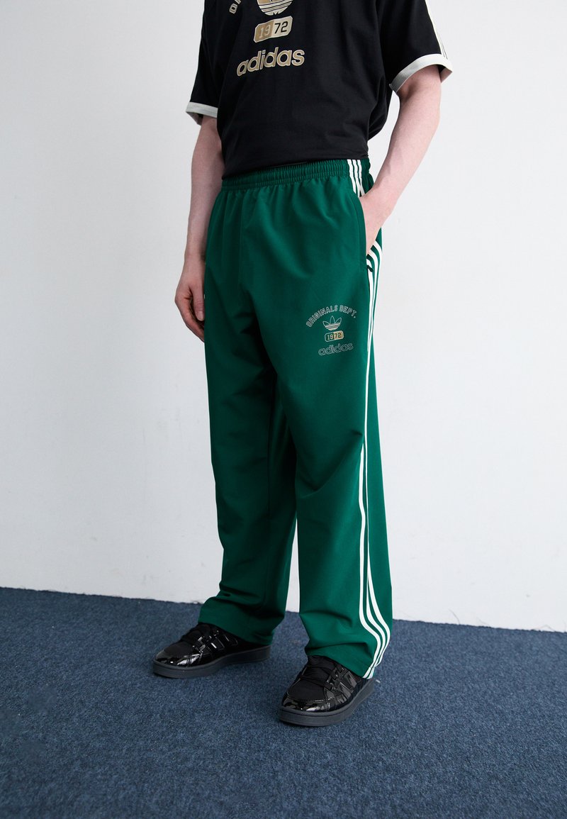 Zelene športne hlače s tremi belimi črtami, z elastičnim pasom in detajlom logotipa, v kombinaciji z črnimi športnimi copati na modrem tepihu.