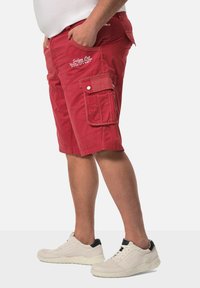 Pantalones cortos cargo rojos con múltiples bolsillos, detalles de costura y un corte relajado. Llevados con zapatillas blancas con acentos negros.
