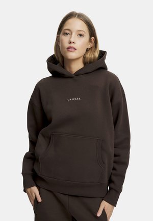Nuori nainen, joka pukeutunut tummanruskeaan huppariin, jossa on etutasku ja "CASPARA"-teksti, seisoo yksivärisen vaalean taustan edessä.